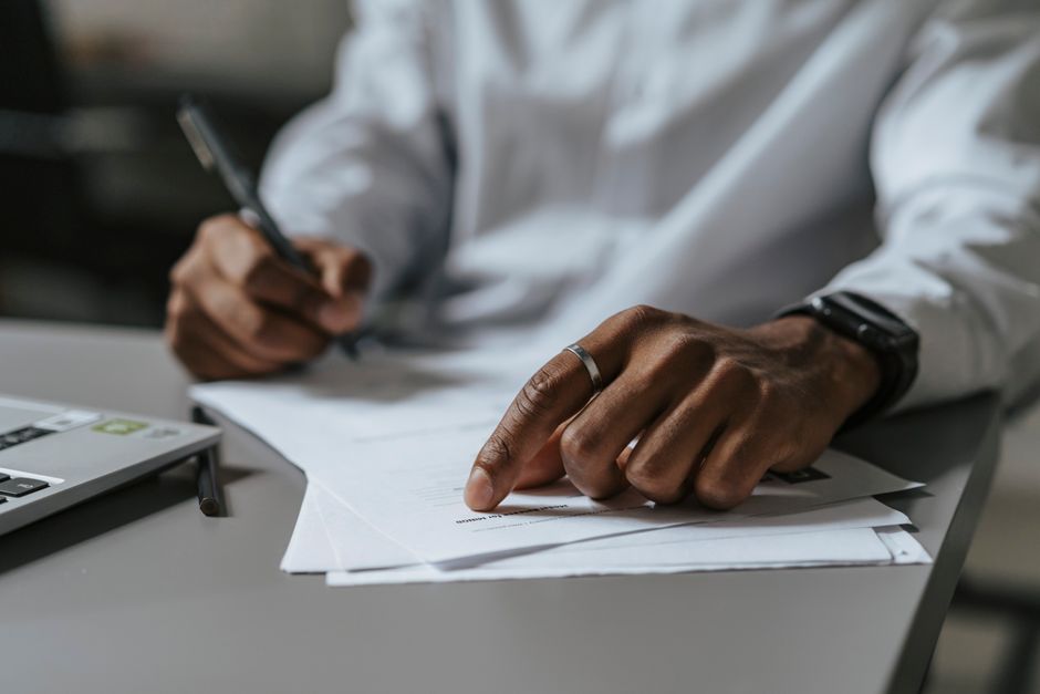 Close-up of a person's hand with a ring, pointing at a document on a light grey table, while holding a pen in the other hand. The individual is wearing a white long-sleeved shirt and a smartwatch. In the background, there is a laptop computer with a partially visible keyboard. The table surface has scattered papers, some of which are being examined or signed, indicating paperwork related to house removals or moving logistics. The scene is indoors, with soft lighting. The context appears to involve formal paperwork or administrative tasks connected to a home relocation, with a focus on the documentation process often associated with house moving services provided by companies like Man with Van North Ockendon.