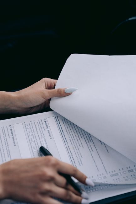 Close-up of a person’s hands holding a partially turned page of a document related to house removals or relocation paperwork, with a black pen held in the other hand. The background is dark and out of focus, emphasizing the paperwork and hands, which are positioned over a table or surface. The scene suggests preparation or organisation involved in a home relocation process, typical of furniture transport or packing and moving activities carried out by Man with Van North Ockendon. The detailed document includes printed text, and the person’s well-manicured nails are visible, indicating attention to detail in the moving planning process.