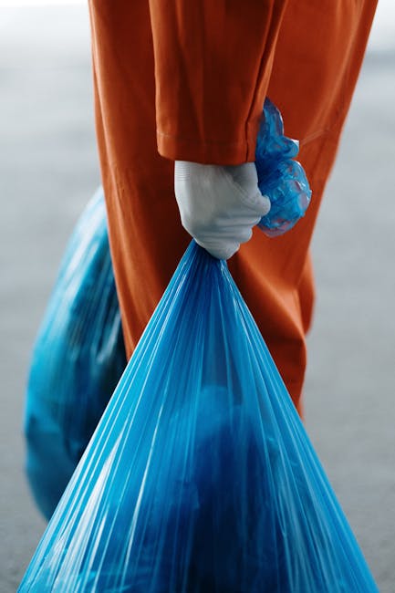 A person wearing an orange protective suit and white gloves is holding a large blue plastic bag filled with waste or recyclable materials. The individual is standing outdoors on a paved or concrete surface, with their arm extended downward, gripping the bag by its gathered top. The background is blurred, emphasizing the focus on the person and the waste collection process. This scene illustrates proper waste handling, which can be associated with home clearance or rubbish removal services typically offered by companies like Man with Van North Ockendon, supporting efficient house removals and relocation logistics.