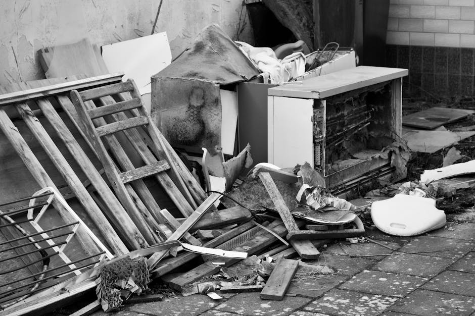 A black-and-white photograph showing a cluttered outdoor area next to a building, featuring a disassembled wooden bed frame leaning against a wall on the left, with wooden slats and broken parts. In front of it, there is a collection of various debris including broken pieces of furniture, cardboard boxes, and plastic items. An old, detached piece of furniture is visible with some fabric and padding exposed, and a white plastic object, possibly a seat or container, is lying on the ground. The ground is paved with square tiles, some of which are damaged or stained. The scene appears to be in a dumping or storage area, likely related to home clearance or waste removal processes. This setting may relate to professional house removals or bulky waste collection services provided by Man with Van North Ockendon as part of their relocation solutions for residential properties.