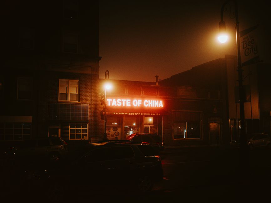 A nighttime exterior view of a brick building housing a restaurant called 'Taste of China,' illuminated by two bright streetlights; the restaurant's red signs are clearly visible, with one displaying its name in bold letters. In front of the building, several parked cars are aligned along the street, and the surrounding area appears quiet. To the right, a street sign indicates parking regulations, and the scene is lit by warm, ambient street lighting, providing enough visibility of the building's facade and nearby pavement. The setting suggests an urban environment suitable for a home relocation process, where moving companies such as Man with Van North Ockendon might coordinate loading or unloading moves with street parking permits during evening hours.