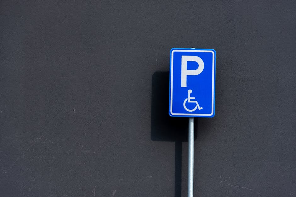 A blue and white parking permit sign indicating accessible parking for disabled individuals is mounted on a metallic pole against a plain dark grey wall. The sign features a large white letter 'P' with a wheelchair symbol below it. The pole is positioned to the right of the image, and the sign casts a subtle shadow on the wall behind it. The surrounding environment suggests an outdoor setting, likely near a property or parking area associated with house removals or relocation services. This type of signage is relevant for home relocation contexts, supporting parking permits for vehicles involved in furniture transport and loading processes, as operated by Man with Van North Ockendon. The muted lighting and minimal background focus attention on the sign itself, providing a clear and precise visual of accessible parking provisions in the area.