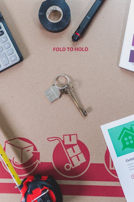 An overhead view of a workspace featuring moving and packing supplies associated with home relocation. Central to the image is a cardboard moving box with red printed instructions, including the words 'FOLD TO HOLD' visible in red ink. Resting on the box are a small silver keychain with a house-shaped charm and a brass key. To the left, a gray calculator is partially visible, and above it, a roll of packing tape with a blue core is placed. To the right of the box, a black marker pen is positioned, and further right, part of a tablet or printed sheet displaying green house icons and informational text can be seen. A yellow tape measure with a red casing is located at the bottom left corner, and a black strap or securing belt is near the top right. The arrangement suggests activities involved in packing, organizing, and preparing items for a house move, typical of services provided by companies like Man with Van North Ockendon within their removals offerings.