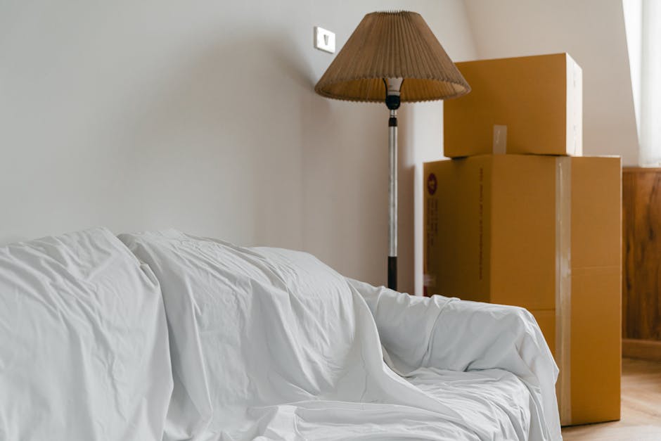 Inside a room during home relocation, several cardboard moving boxes are stacked against a light-colored wall near a window, with sunlight illuminating the area. A wooden cabinet is partially visible in the background. In the foreground, a white sofa covered with a slipcover is positioned close to the boxes. To the left of the boxes, a tall floor lamp with a brown fabric shade stands on the wooden floor, providing additional lighting. The scene depicts the process of packing and preparing for furniture transport, with visible packaging materials such as cardboard boxes and the arrangement of furniture and belongings in a typical domestic setting, indicating a relocation or moving process facilitated by Man with Van North Ockendon.
