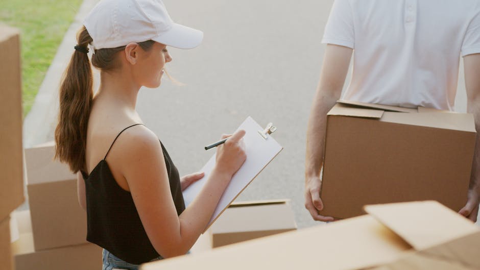An overhead view of a workspace featuring moving and packing supplies associated with home relocation. Central to the image is a cardboard moving box with red printed instructions, including the words 'FOLD TO HOLD' visible in red ink. Resting on the box are a small silver keychain with a house-shaped charm and a brass key. To the left, a gray calculator is partially visible, and above it, a roll of packing tape with a blue core is placed. To the right of the box, a black marker pen is positioned, and further right, part of a tablet or printed sheet displaying green house icons and informational text can be seen. A yellow tape measure with a red casing is located at the bottom left corner, and a black strap or securing belt is near the top right. The arrangement suggests activities involved in packing, organizing, and preparing items for a house move, typical of services provided by companies like Man with Van North Ockendon within their removals offerings.
