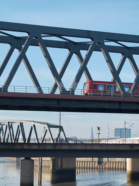 A red passenger tram moving along an elevated railway bridge with a modern, steel truss structure. The bridge spans over a river, with a second, lower railway bridge visible beneath it. The scene is set against a clear blue sky, with the river visible below and the city skyline with high-rise buildings in the background. The image depicts a transportation infrastructure likely involved in urban travel and commuting, relevant to logistical considerations in house removals and home relocation services offered by Man with Van North Ockendon, especially when planning routes near Rainham Marshes.
