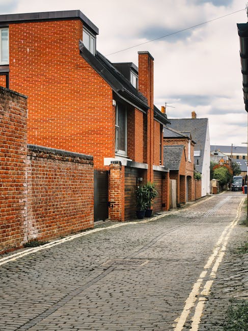 A narrow paved driveway in North Ockendon lined with red brick residential buildings on one side and an old brick wall on the other. The buildings have multiple levels with windows, some with black frames, and small front doors. A large potted plant is visible near one of the entrances. Overhead, cloudy skies cast diffuse light on the scene. The driveway is used for access and parking, with a vehicle visible in the distance. This setting illustrates the type of environment where house removals and furniture transport by Man with Van North Ockendon are often carried out, especially in tight urban or suburban areas requiring careful planning for loading and unloading during home relocation or packing and moving projects.