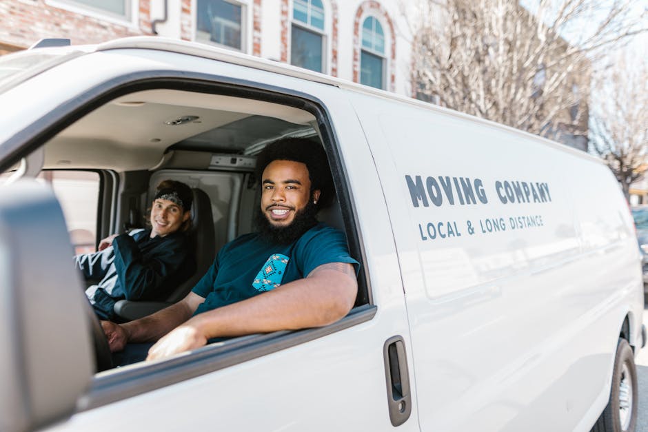 Inside the cab of a white moving van from Man with Van North Ockendon, two movers are visible. The driver, a man with dark curly hair and a beard, is smiling at the camera, wearing a dark blue t-shirt