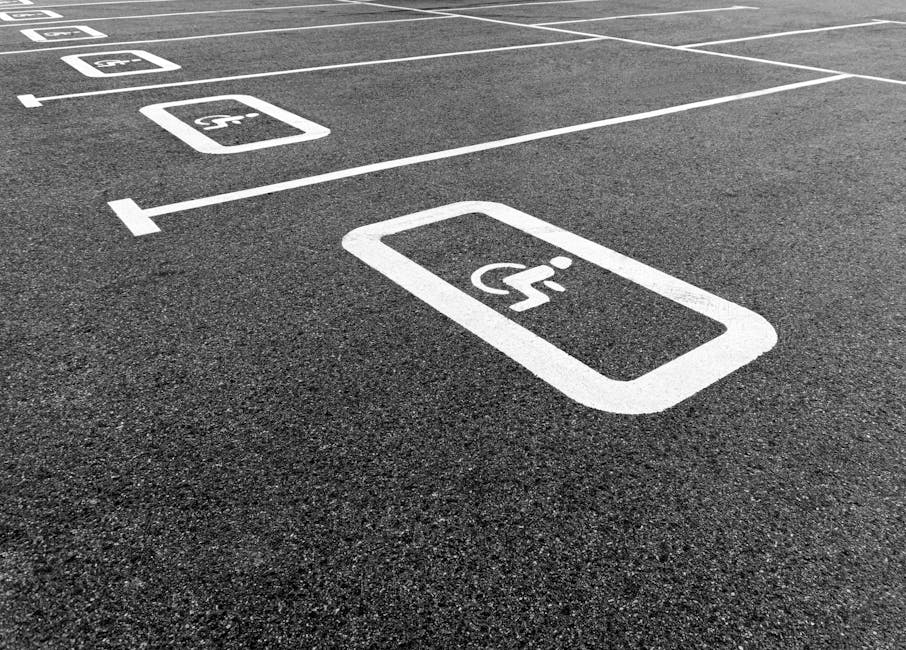 Close-up view of a parking lot pavement with white painted parking space lines, specifically highlighting designated accessible parking bays marked with wheelchair symbols inside rectangles. The markings are crisp and clearly visible on the dark asphalt surface, which has a rough texture. The parking spaces are aligned in a row, with some spaces showing clearly in the foreground and others extending into the background. These designated accessible parking spots are positioned near the entrance of a property, which is relevant for home relocation and furniture transport services. The image suggests an organized loading area suitable for professional removals, such as those provided by Man with Van North Ockendon, supporting the ease of vehicle access during moving operations.