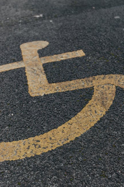 Close-up view of a disabled parking space marking on tarmac pavement, featuring a yellow wheelchair symbol painted with slightly worn edges. In the background, a white van with an open rear doorway is partially visible, positioned on the pavement near the marked space. Inside the van, several cardboard boxes and packing materials are stacked, with some wrapped in plastic or fabric covers. Two movers, partially visible, are seen lifting or carrying a wooden furniture piece toward the van, indicating a home relocation process. The scene takes place outdoors during daylight, with natural lighting highlighting the textures of the asphalt, the painted symbol, and the packing materials. The image supports themes of furniture transport, packing, and loading during house removals, aligning with professional moving services offered by Man with Van North Ockendon, specifically for house removals and moving logistics.