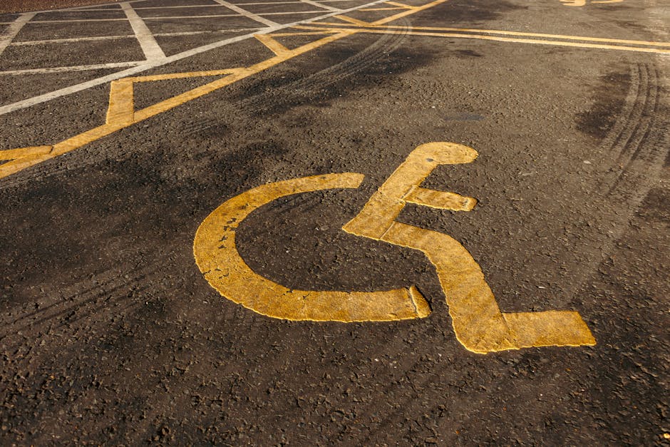 Close-up view of a parking lot pavement with white painted parking space lines, specifically highlighting designated accessible parking bays marked with wheelchair symbols inside rectangles. The markings are crisp and clearly visible on the dark asphalt surface, which has a rough texture. The parking spaces are aligned in a row, with some spaces showing clearly in the foreground and others extending into the background. These designated accessible parking spots are positioned near the entrance of a property, which is relevant for home relocation and furniture transport services. The image suggests an organized loading area suitable for professional removals, such as those provided by Man with Van North Ockendon, supporting the ease of vehicle access during moving operations.